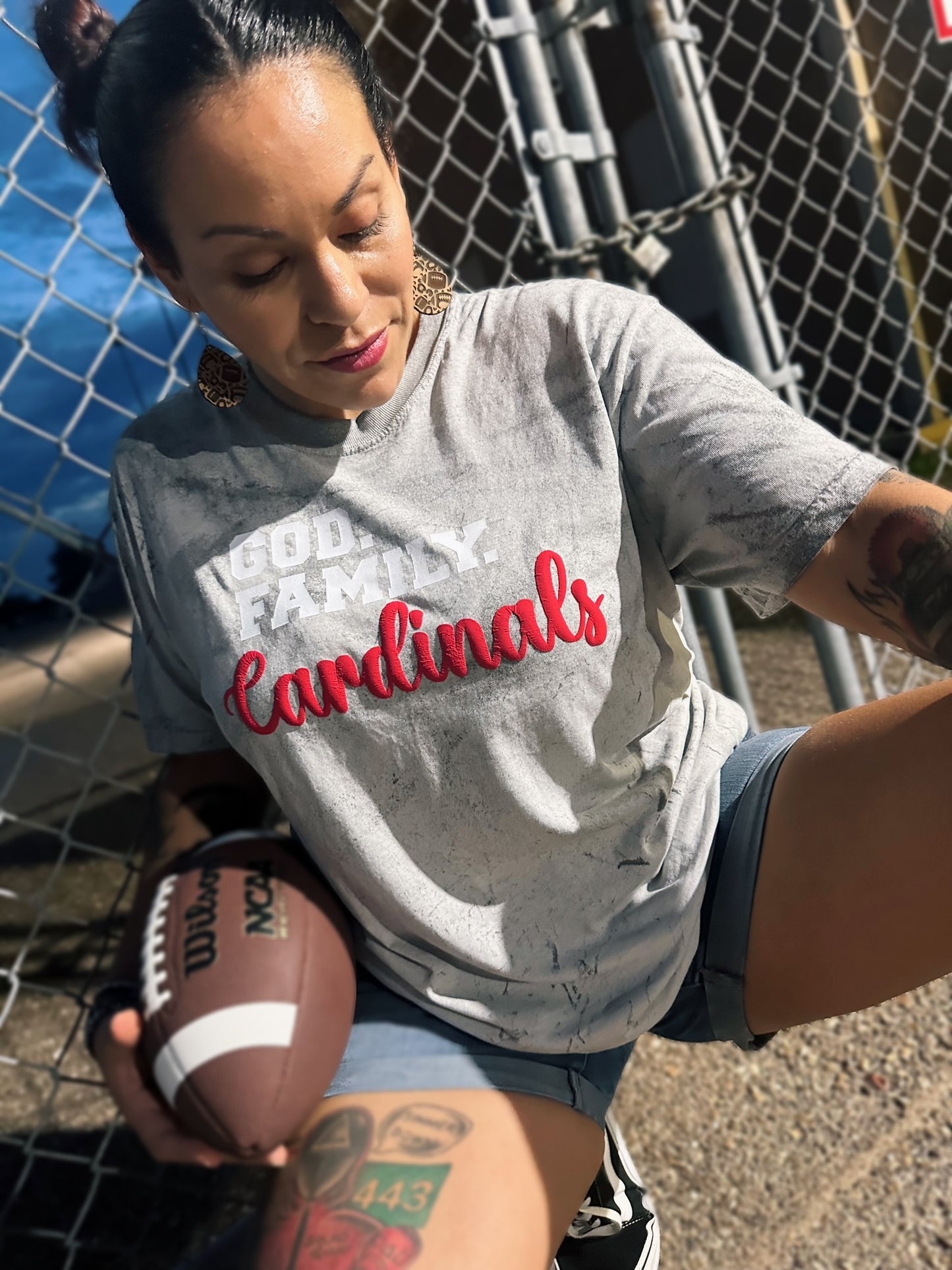 Woman wearing a 'Cardinals' shirt holding a football, standing in front of a chain-link fence.