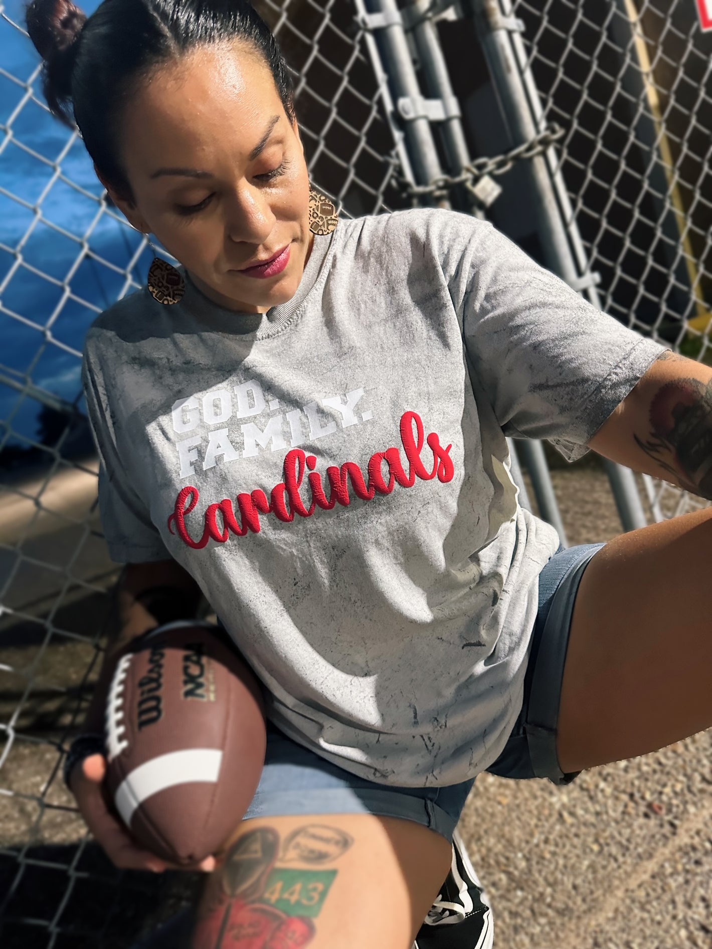 Woman wearing a 'Cardinals' shirt holding a football, standing in front of a chain-link fence.